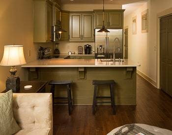 Green cabinets on line the walls in the kitchen with an  L shape counter with two wooden stools. Medium wood floors 